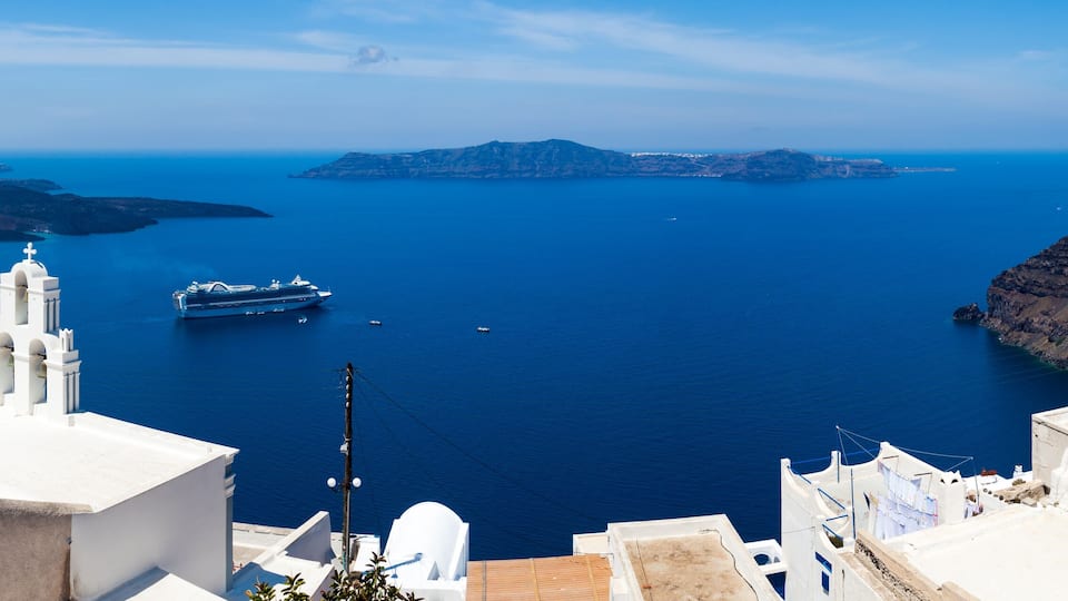 A coastal town with white buildings and blue domes, a large cruise ship, and a clear view of the surrounding islands and sea.