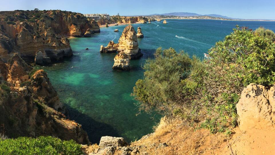 A coastal landscape with rocky cliffs, clear turquoise waters, and distant buildings.