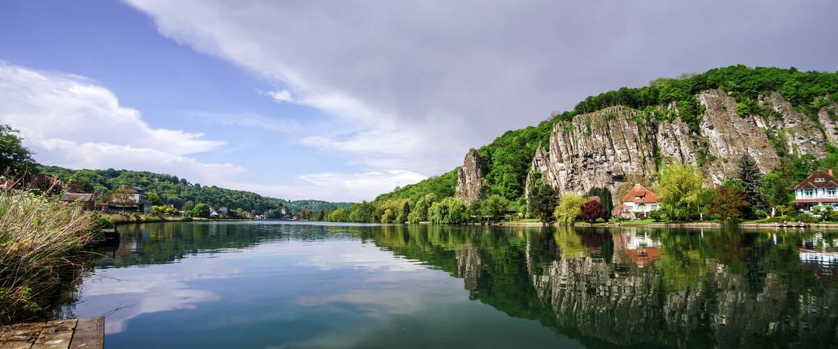 A calm river with a wooden dock, houses, and a cliff with greenery.