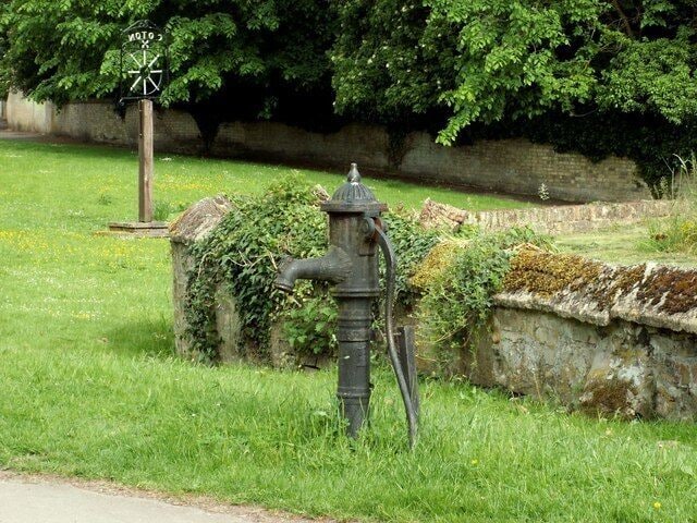 Village pump at Coton This pump stands by the parish church and the low wall forms part of the graveyard boundary. The village sign can be seen in background.