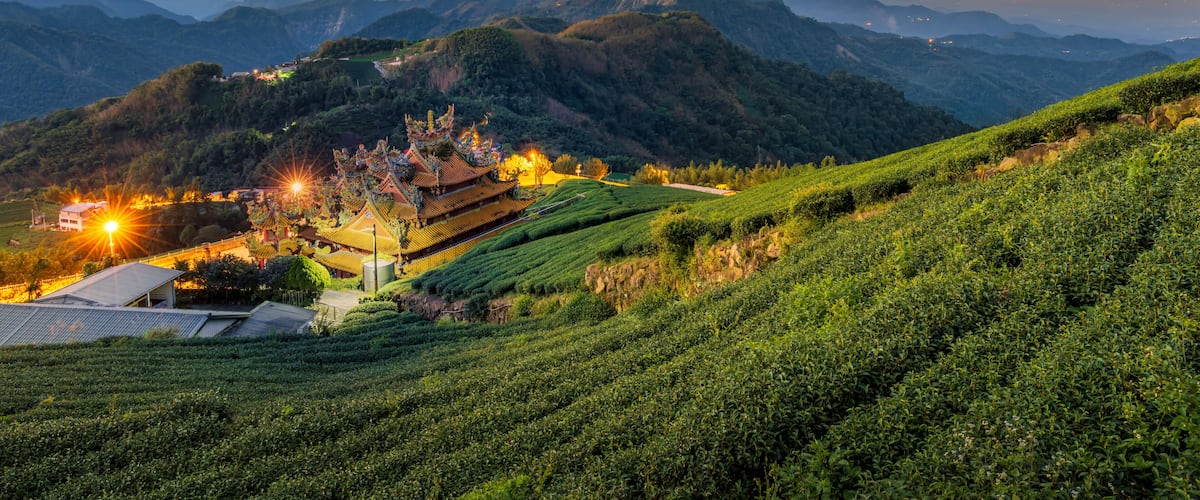 A traditional temple complex nestled in a lush valley with terraced fields and distant mountains under a twilight sky.