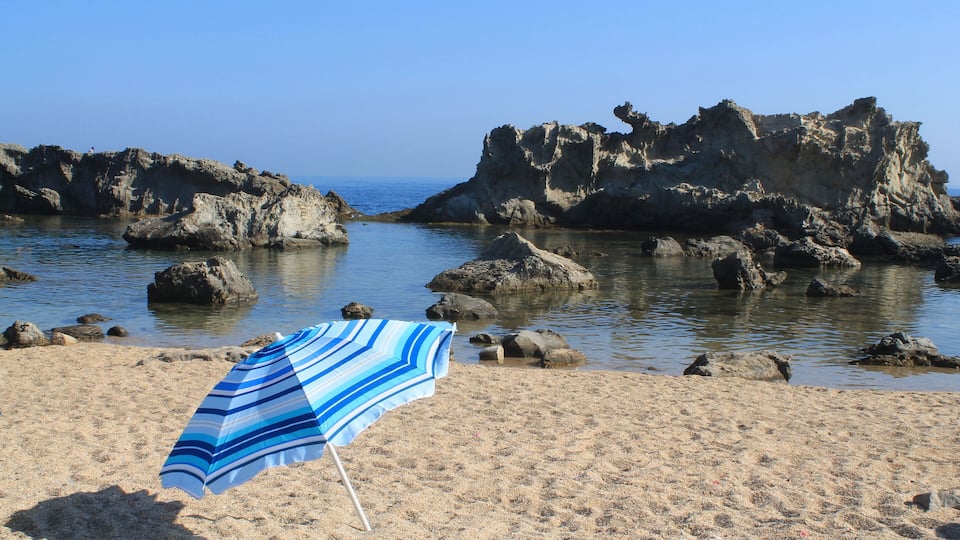 A beach with a striped beach umbrella, rocky formations, and calm water.