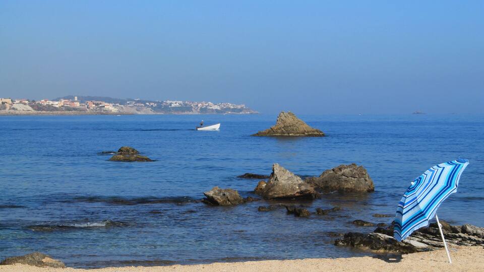 A beach with a striped beach umbrella, rocks, and a small boat in the water.