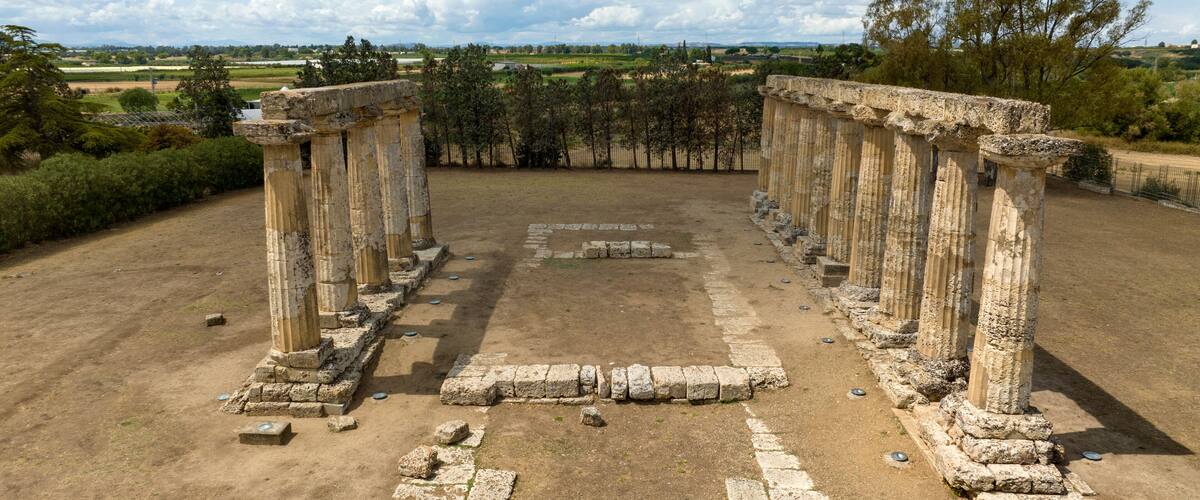 Ancient ruins with columns and stone blocks, surrounded by a rural landscape.