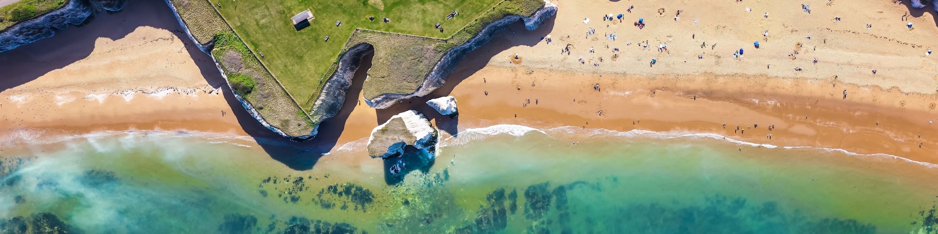 Une vue aĂ©rienne dâune plage aux eaux cristallines, avec une zone herbeuse et un parking.