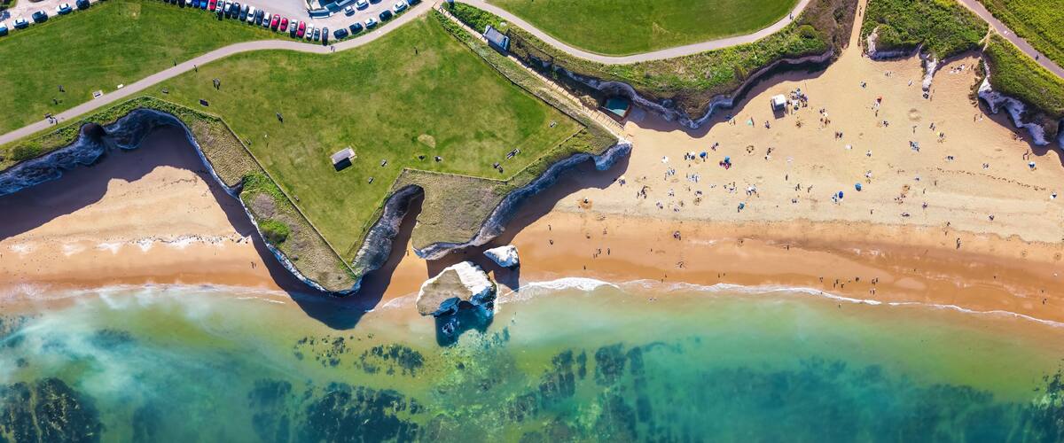 An aerial view of a beach with clear waters, a grassy area, and a parking lot.