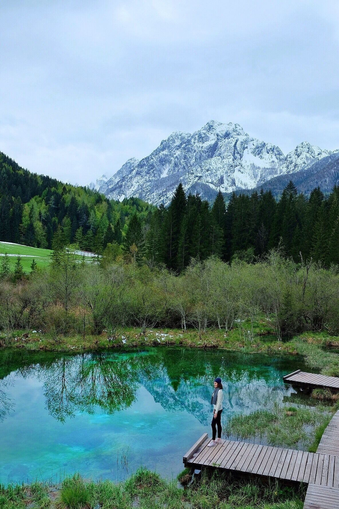 A person standing on a wooden dock by a clear blue lake with snow-capped mountains in the background.