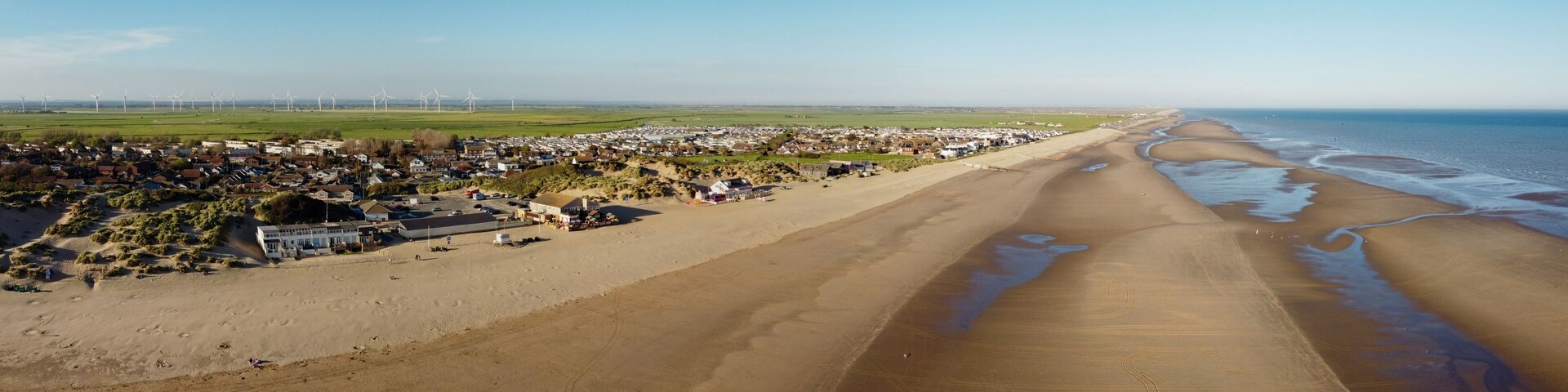 A coastal town with a beach, houses, and wind turbines in the distance.