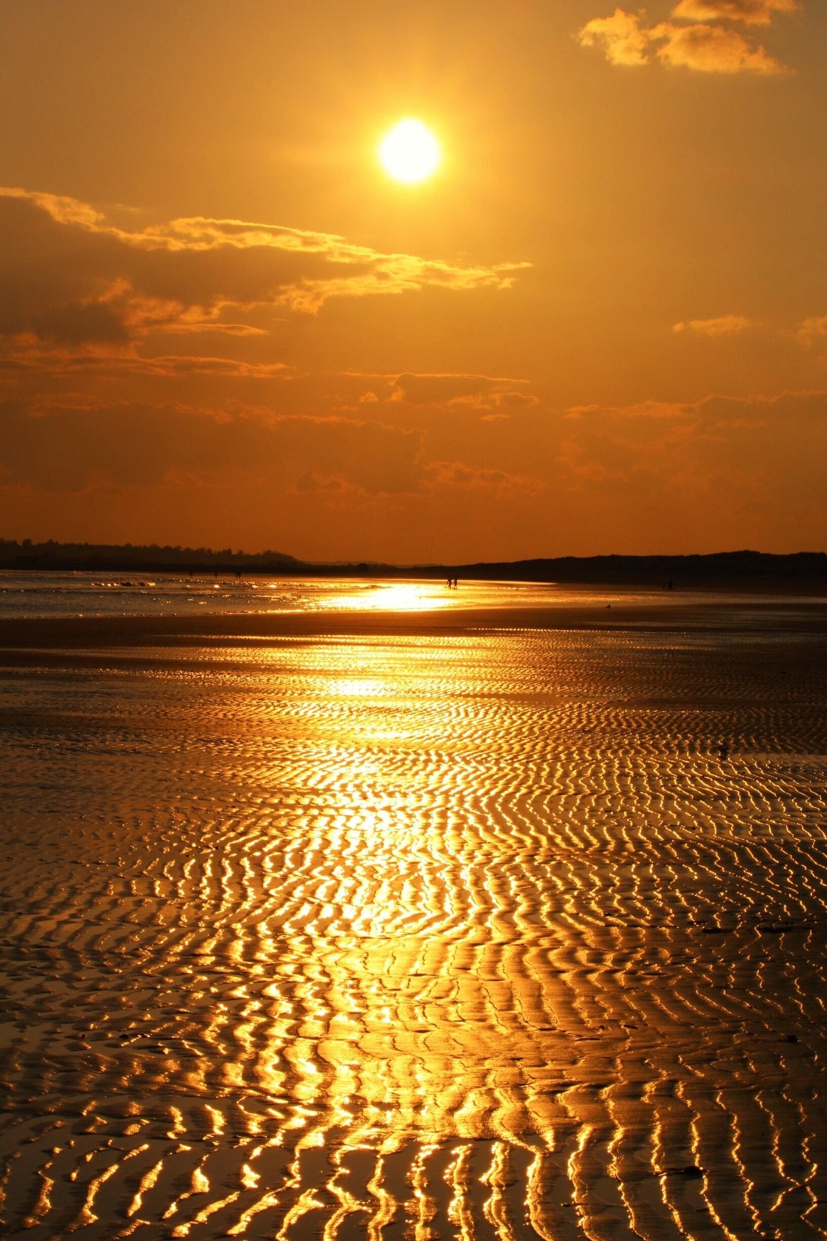 A sunset over a beach with rippled sand patterns and distant silhouettes of people.
