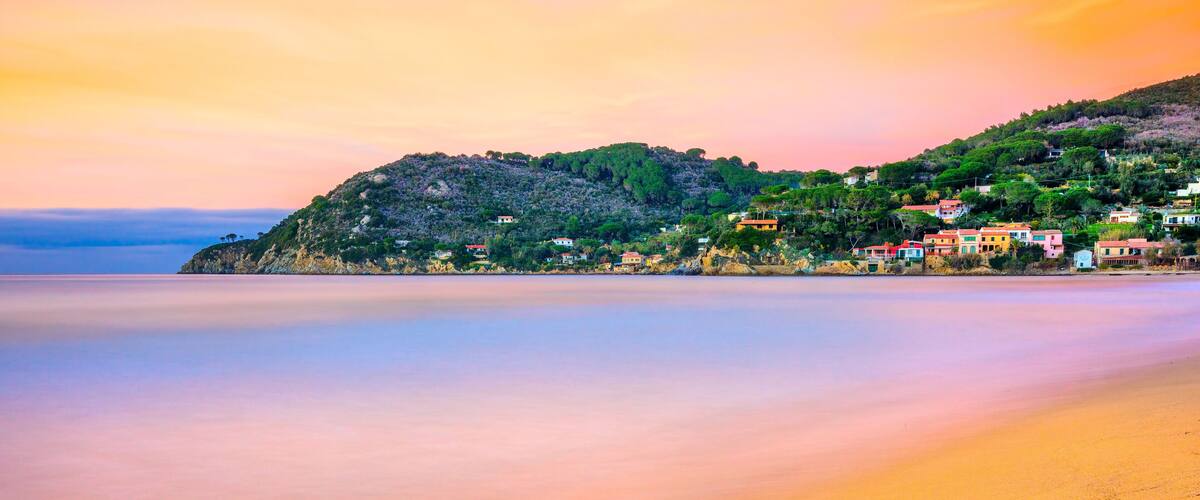 A coastal landscape with a beach, a hill with houses, and a calm sea.