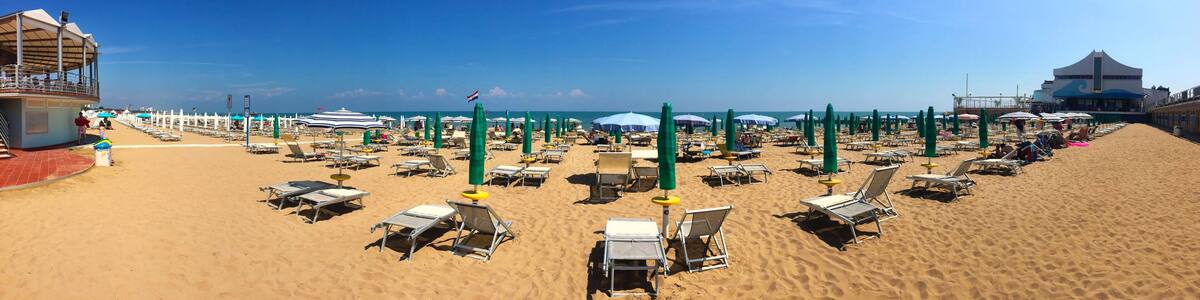 Une plage bordée de rangées de chaises longues et de parasols, une tour de surveillance pour les sauveteurs, et un bâtiment dont la façade est ornée de motifs évoquant les vagues.