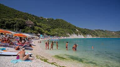 A crowded beach with people sunbathing and swimming, surrounded by lush green hills.