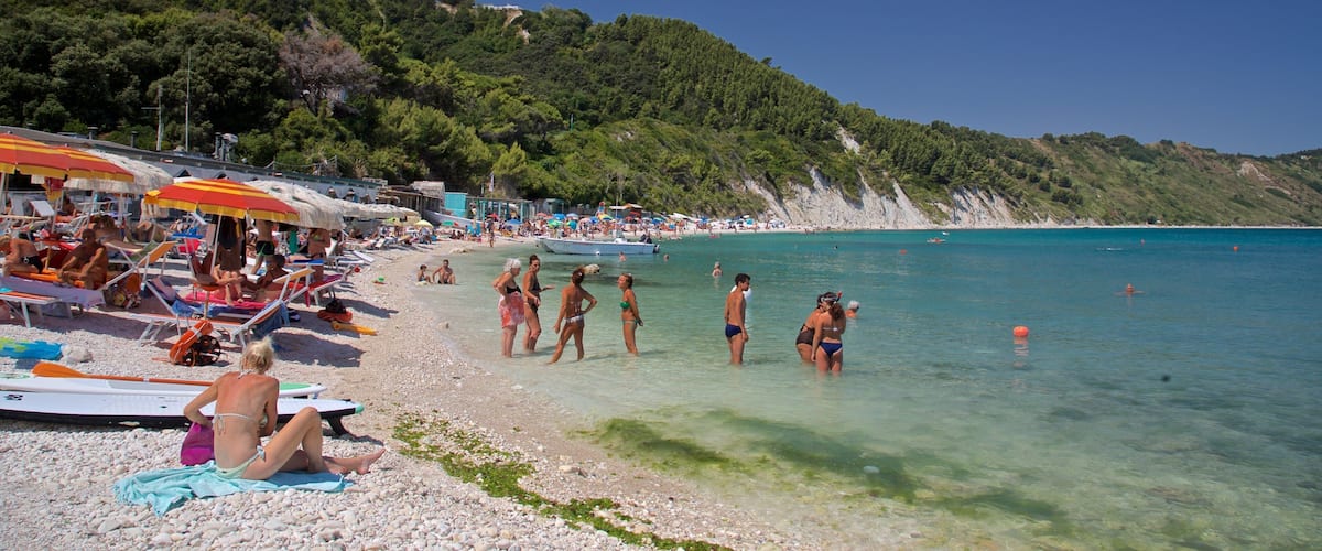 A crowded beach with people sunbathing and swimming, surrounded by lush green hills.
