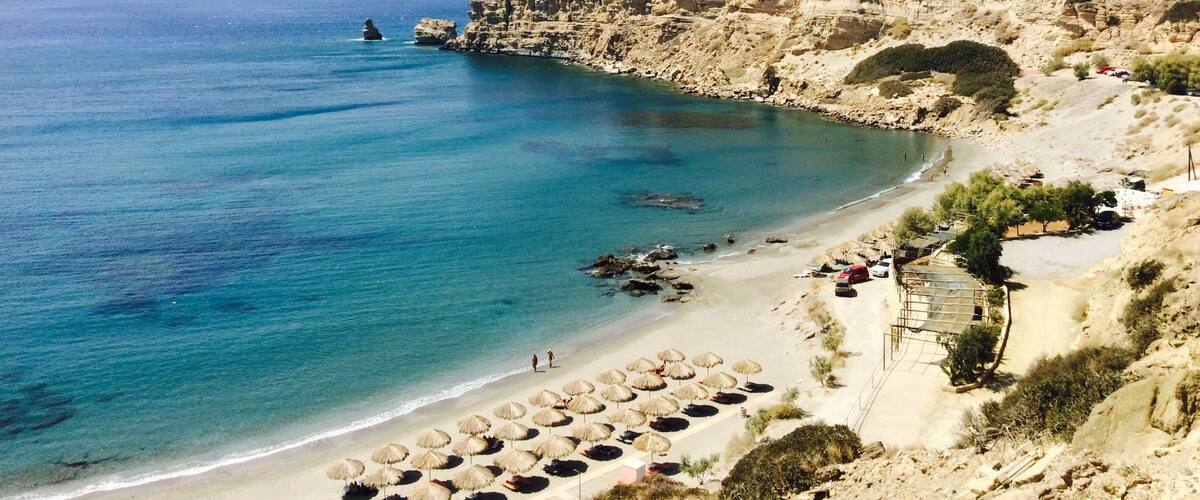A coastal landscape with a rocky cliff, a beach with umbrellas, and a clear blue sea.