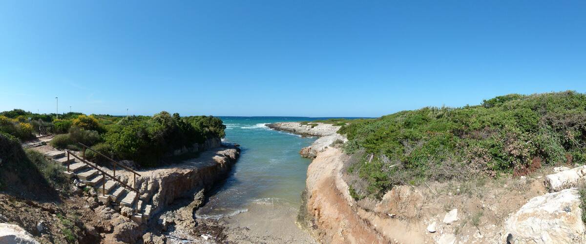 A coastal landscape with a narrow waterway, rocky terrain, and dense vegetation.