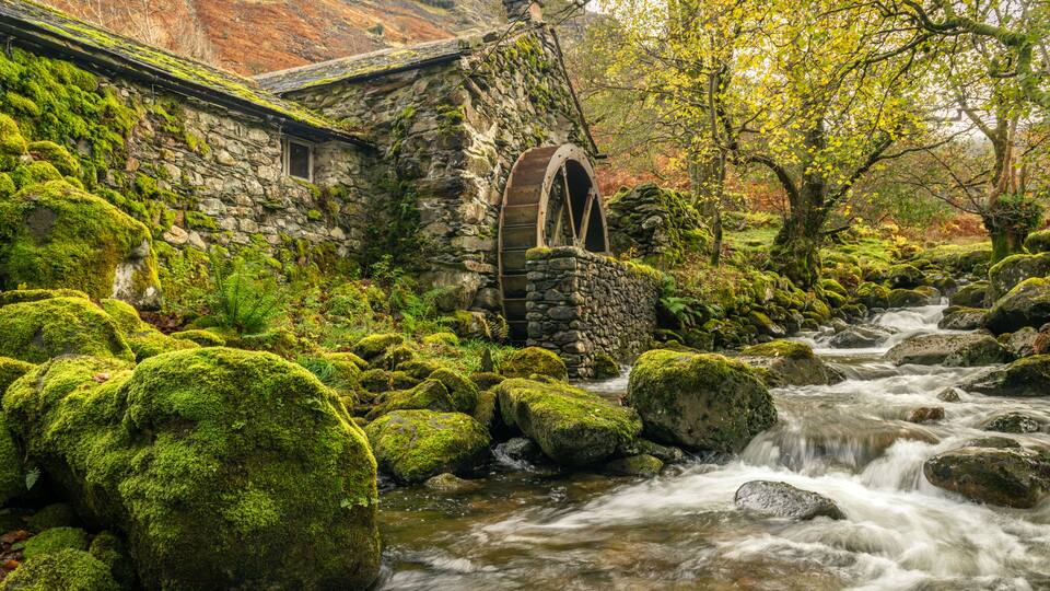A stone building with a moss-covered roof, a wooden water wheel, and a flowing stream with moss-covered rocks.