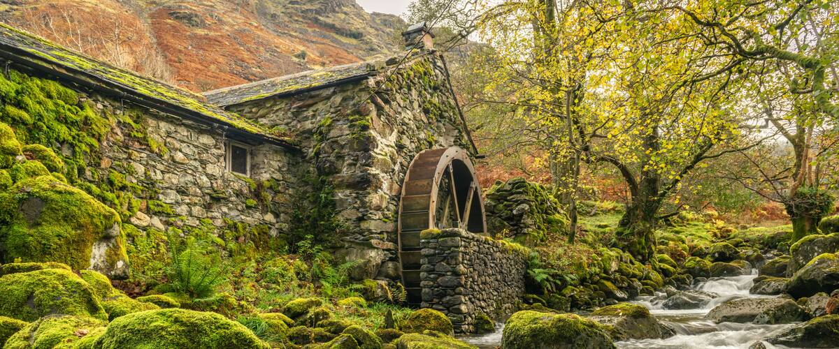 A stone building with a moss-covered roof, a wooden water wheel, and a flowing stream with moss-covered rocks.