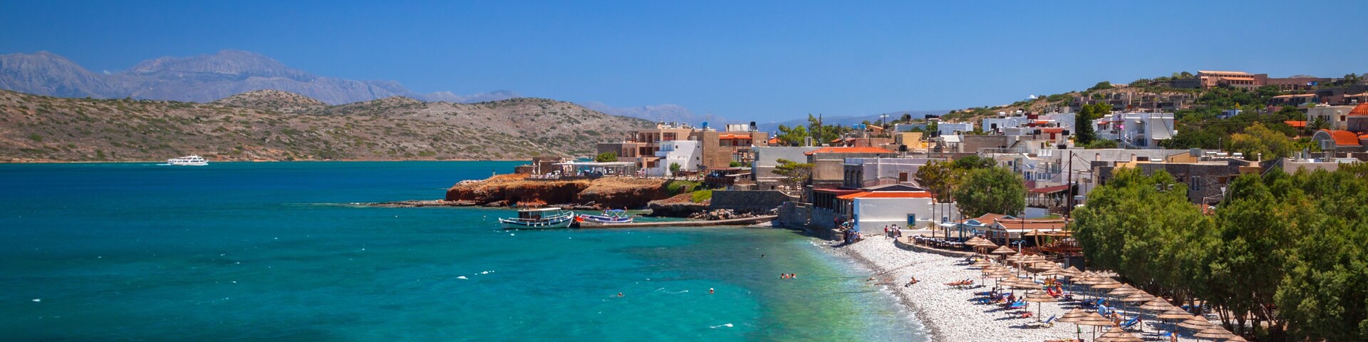 Un pueblo costero con mar azul cristalino, una playa de arena con sombrillas y montañas al fondo.