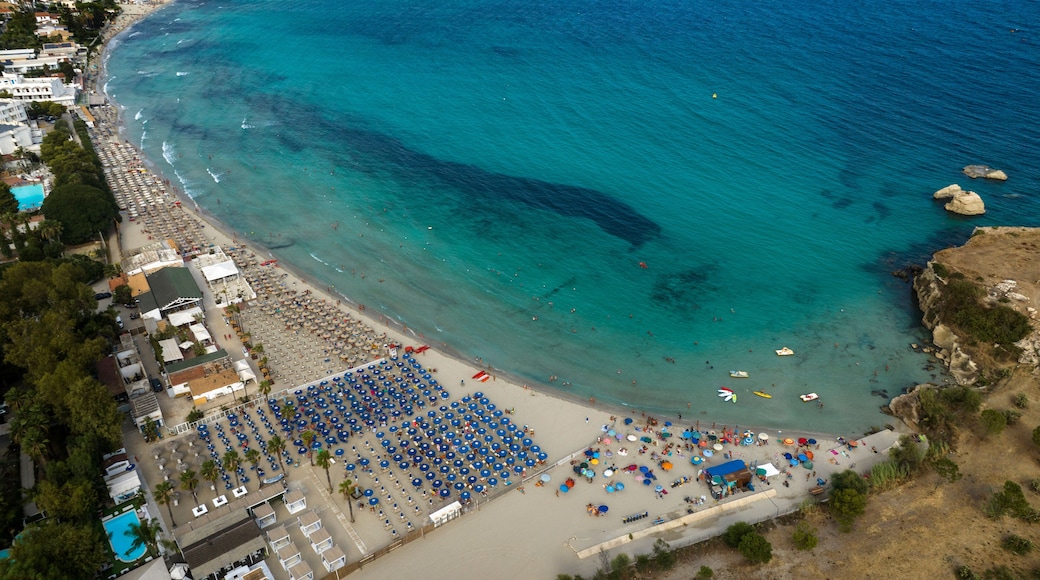Vista aérea de uma vila costeira com uma praia, águas turquesa cristalinas e diversos guarda-sóis e espreguiçadeiras.
