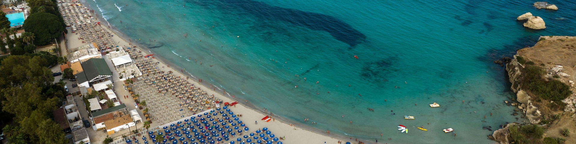 An aerial view of a coastal town with a beach, clear turquoise waters, and a variety of beach umbrellas and sun loungers.