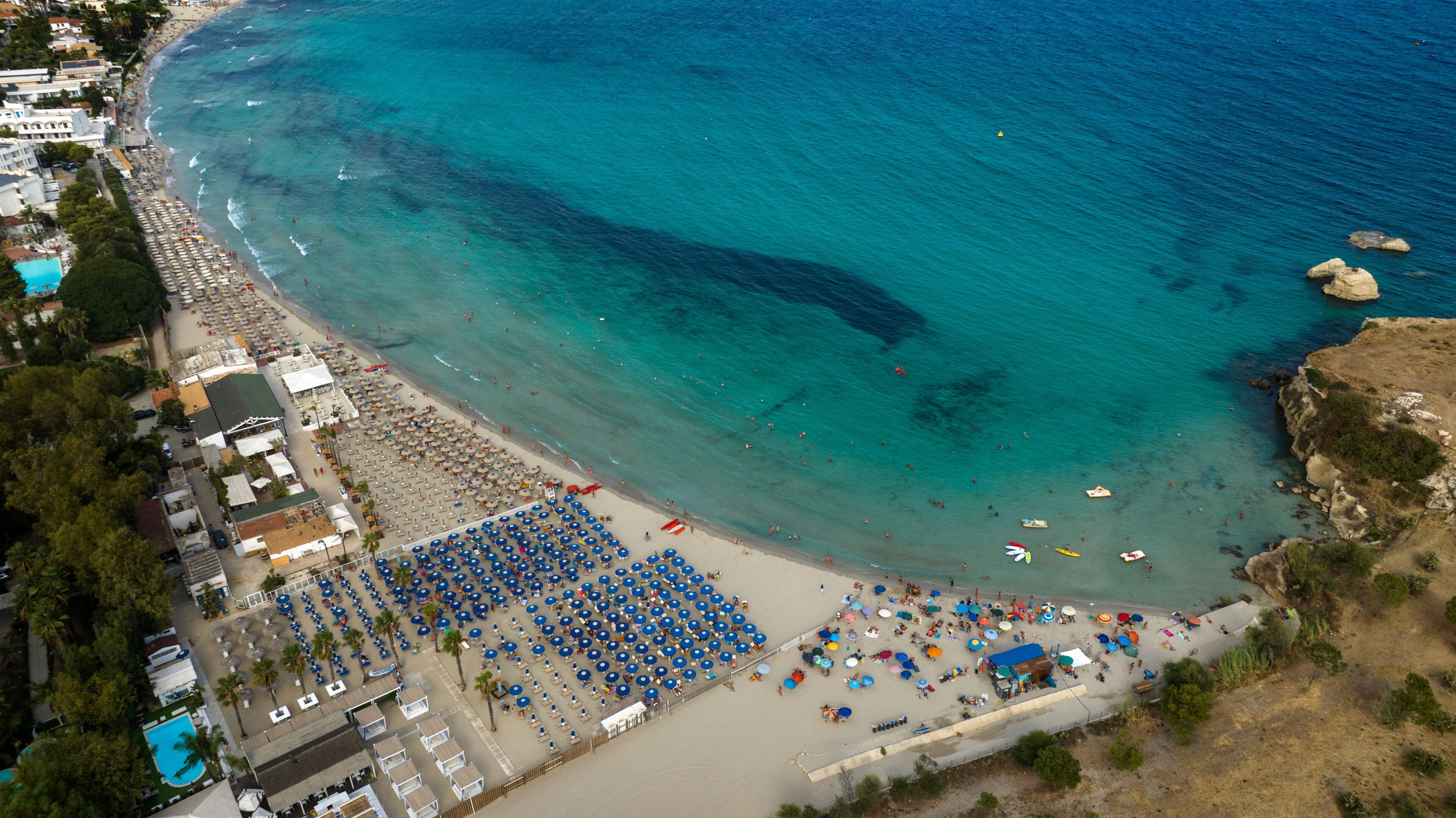 An aerial view of a coastal town with a beach, clear turquoise waters, and a variety of beach umbrellas and sun loungers.