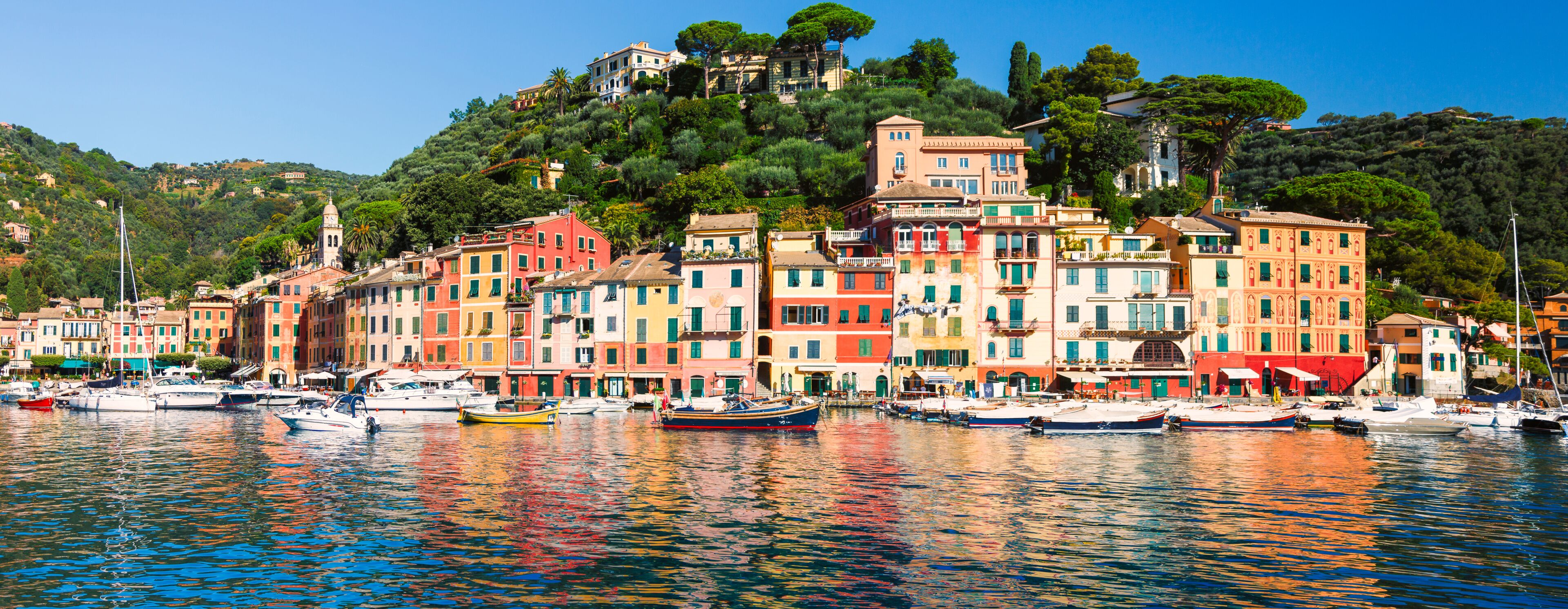 A coastal town with colorful buildings, boats docked along the waterfront, and a hill with trees in the background.