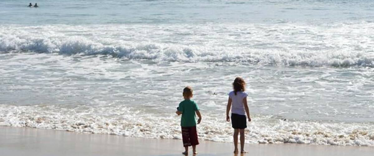 Two people walking on a beach with waves crashing onto the shore and a distant shoreline with vegetation.