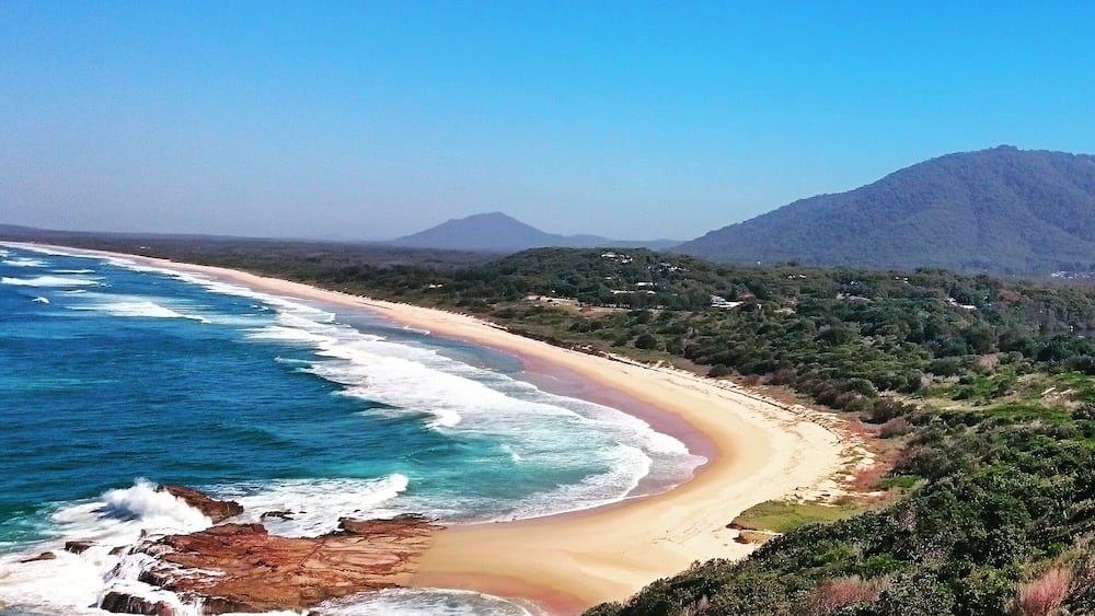 Eine Küstenlandschaft mit Sandstrand, Wellen im Meer und einem Berg im Hintergrund.