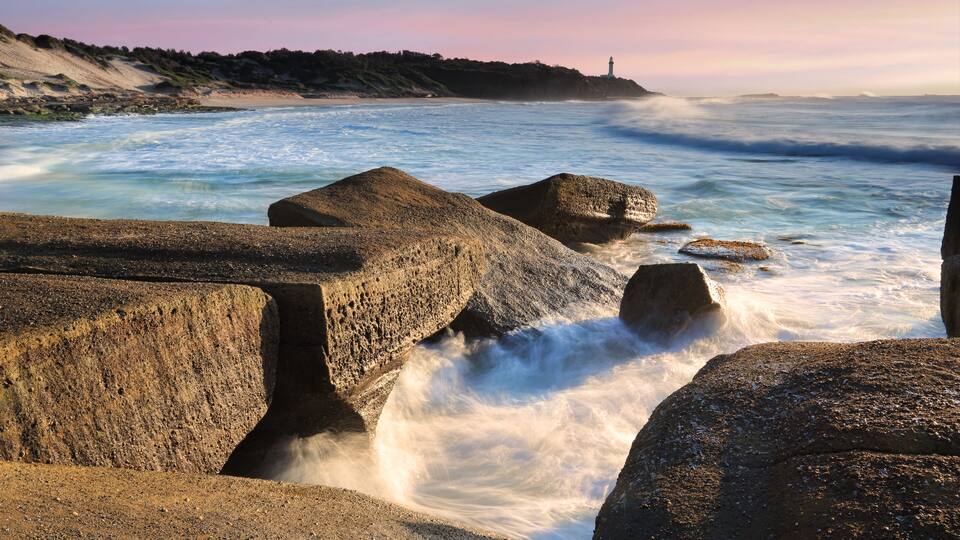 A rocky coastline with waves crashing against the rocks and a lighthouse in the distance.