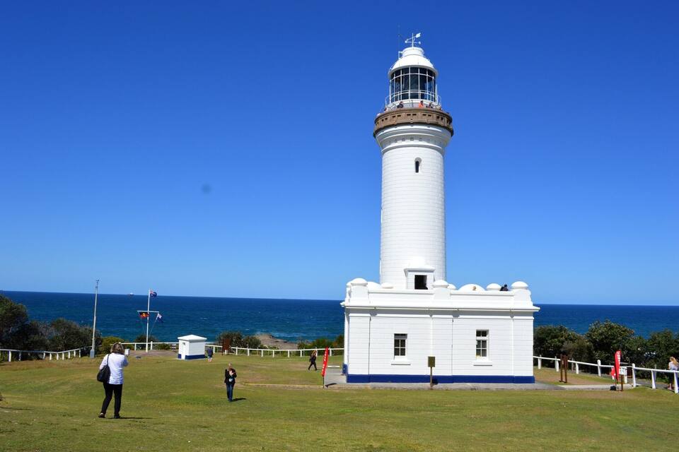 A white lighthouse with a blue base, surrounded by a grassy area and a white fence, with the ocean in the background.