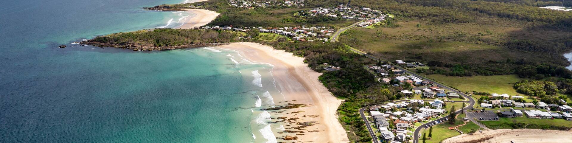 A coastal landscape with a sandy beach, clear turquoise waters, and a residential area with houses and roads.