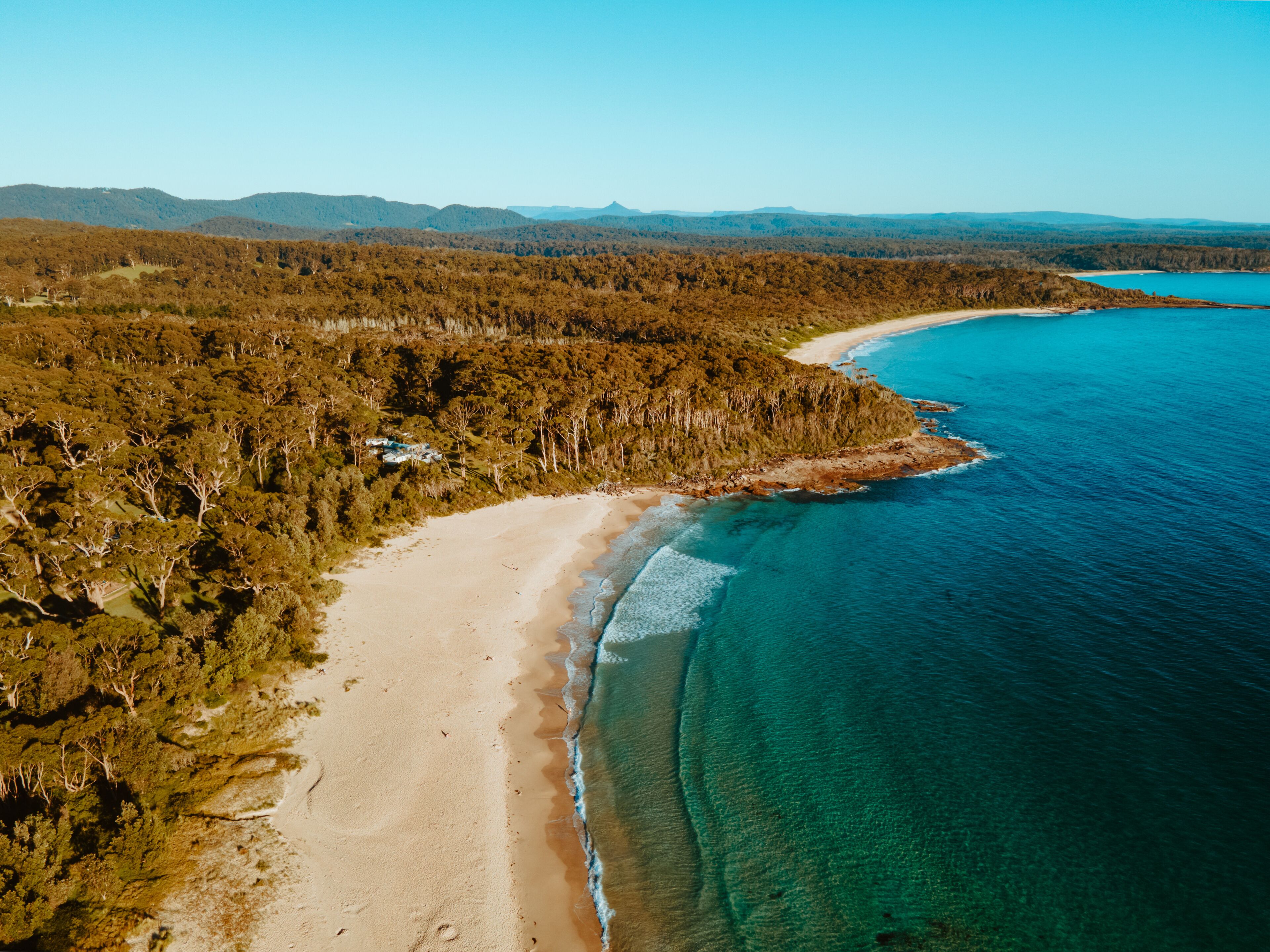 Eine Küstenlandschaft mit Sandstrand, klarem, blauem Wasser und bewaldeten Hügeln im Hintergrund.