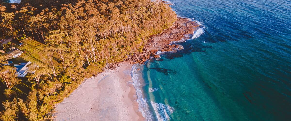 An aerial view of a beach with clear blue waters, sandy shore, and dense forested area.