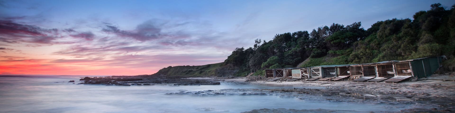 A beach with a sunset, rocky shoreline, and a row of wooden structures.