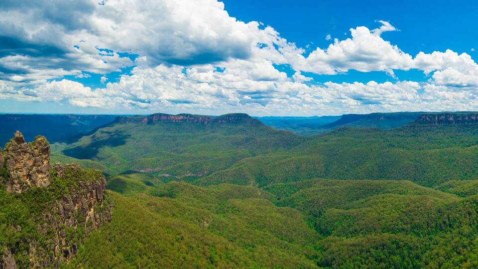 A mountainous landscape with dense green forests and rocky cliffs under a partly cloudy sky.