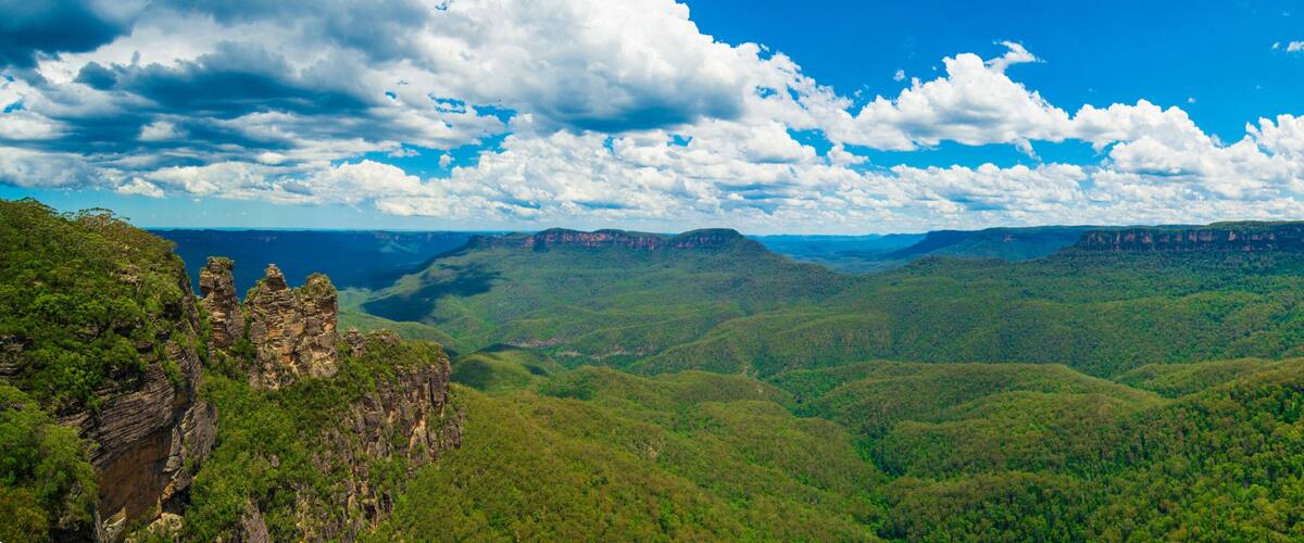 A mountainous landscape with dense green forests and rocky cliffs under a partly cloudy sky.