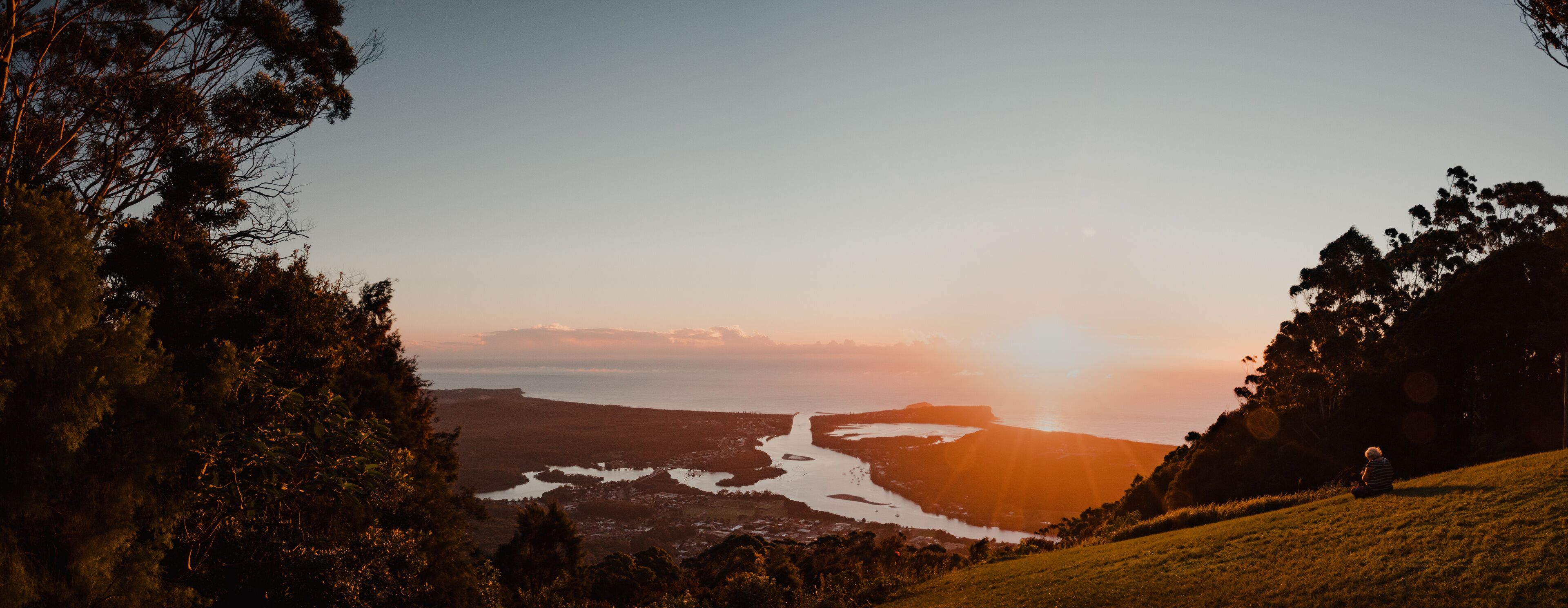 A coastal landscape with a river, hills, and a person sitting on a grassy hill during sunset.
