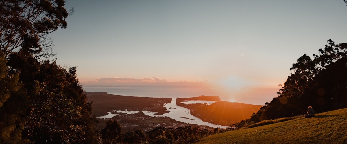 A coastal landscape with a river, hills, and a person sitting on a grassy hill during sunset.