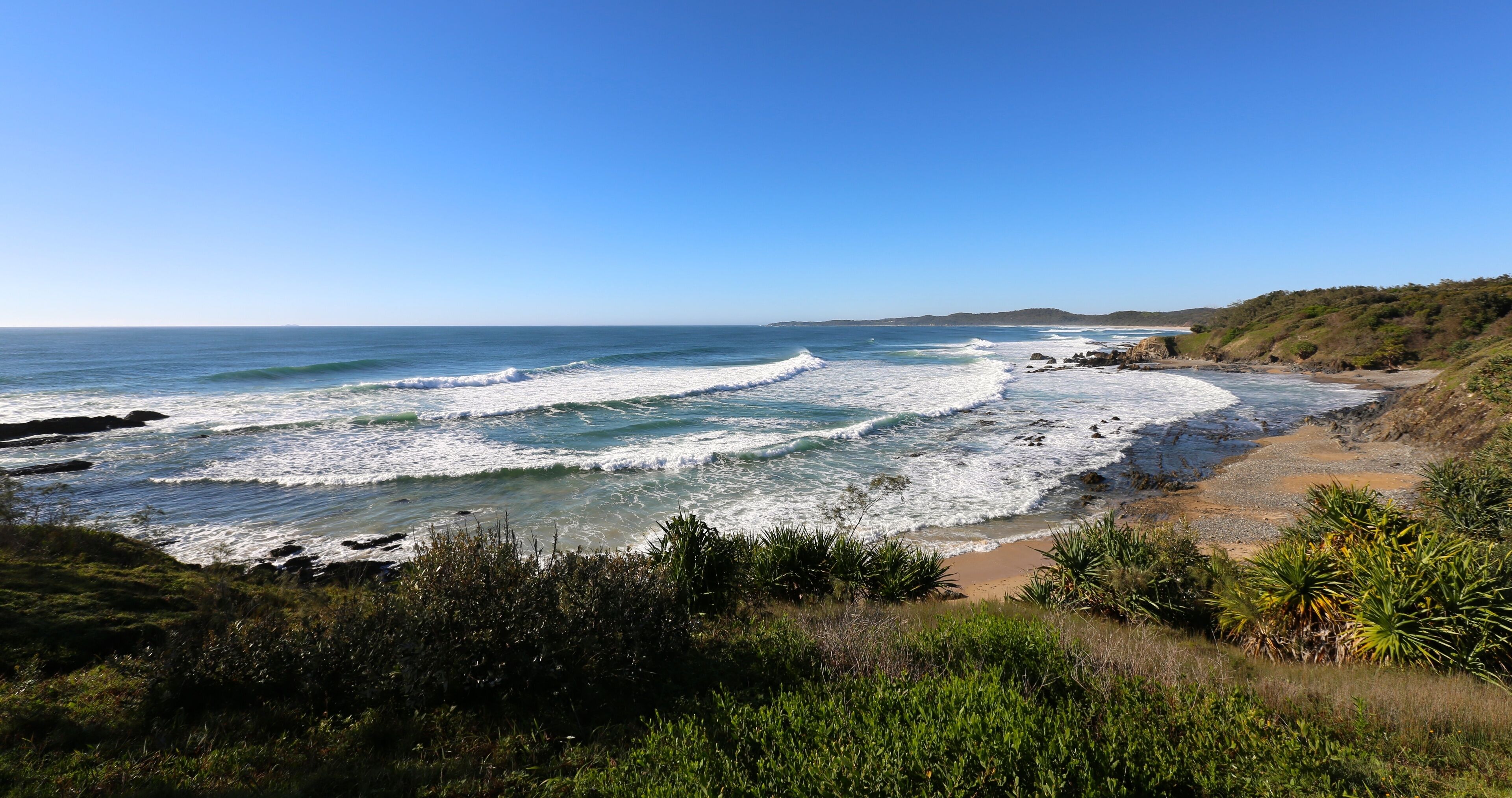 A coastal landscape with waves crashing onto a sandy beach, surrounded by greenery and hills.