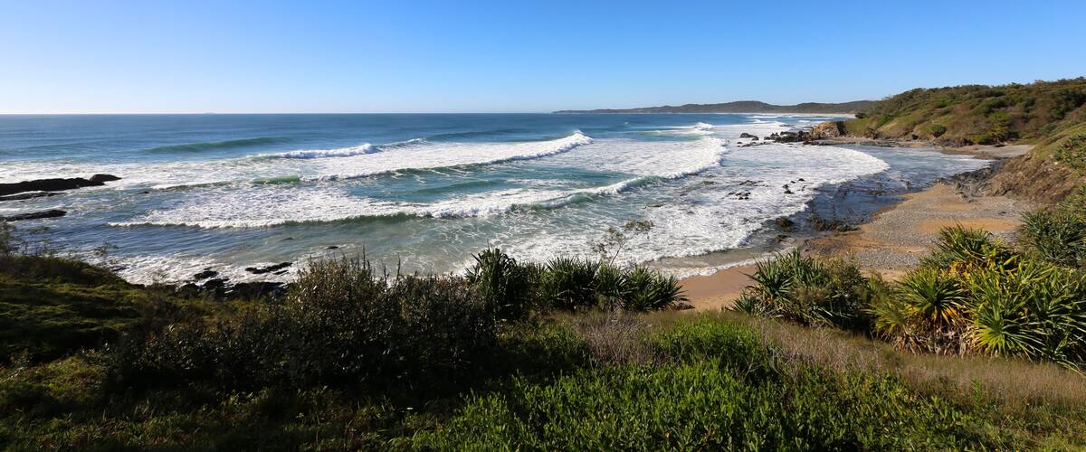 A coastal landscape with waves crashing onto a sandy beach, surrounded by greenery and hills.