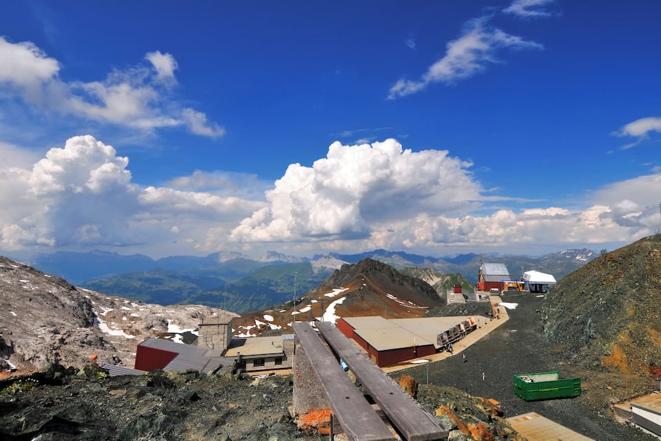 Un paysage montagneux avec un ciel dégagé, des zones enneigées et des bâtiments.