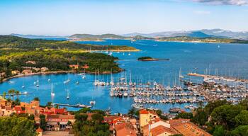 Uma cidade costeira com um porto cheio de veleiros, rodeada por vegetação exuberante e colinas ao fundo.