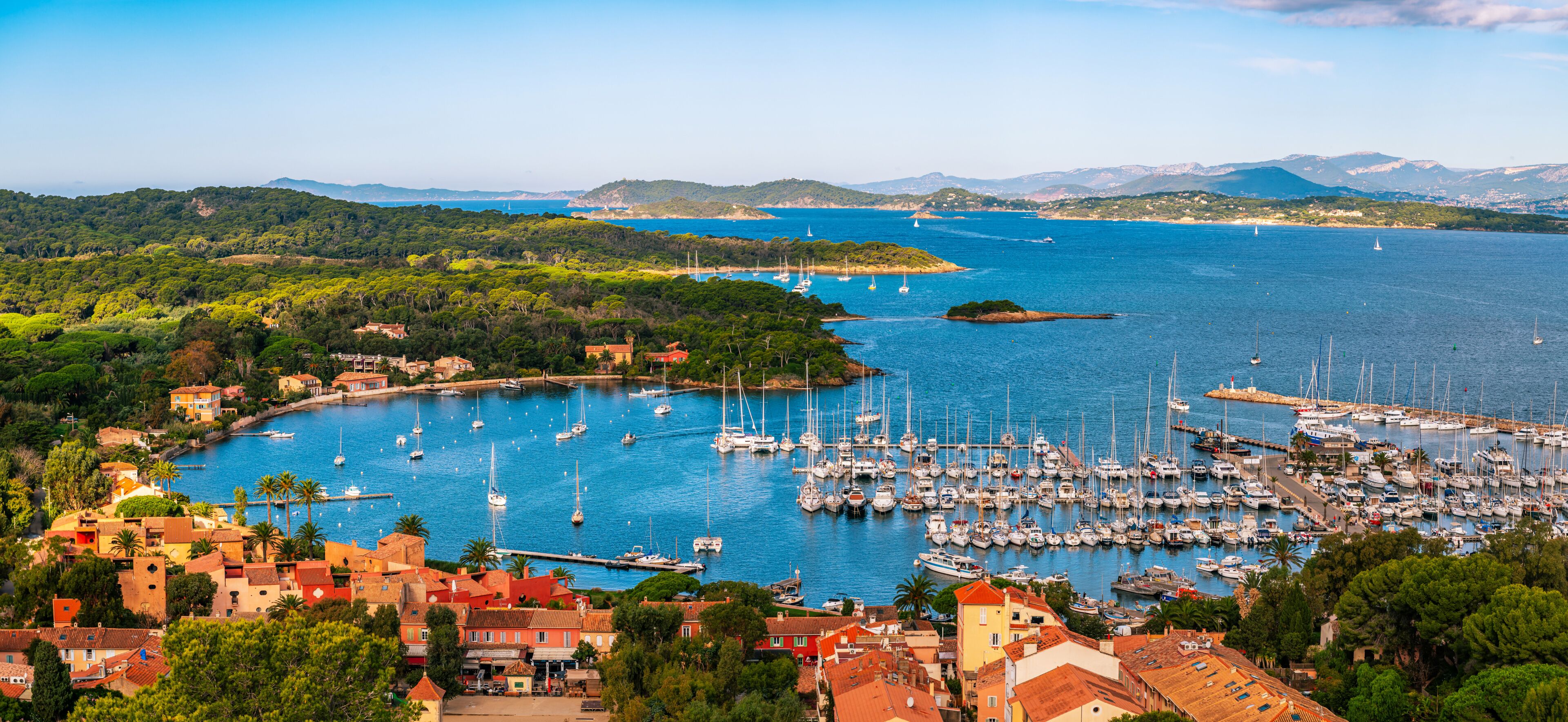 A coastal town with a marina filled with sailboats, surrounded by lush greenery and hills in the background.