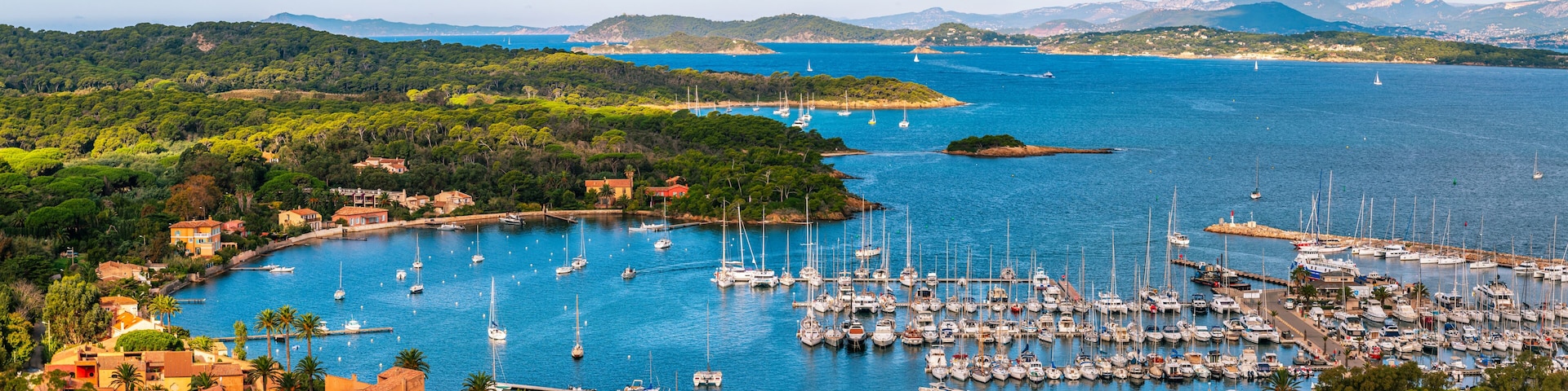 A coastal town with a marina filled with sailboats, surrounded by lush greenery and hills in the background.