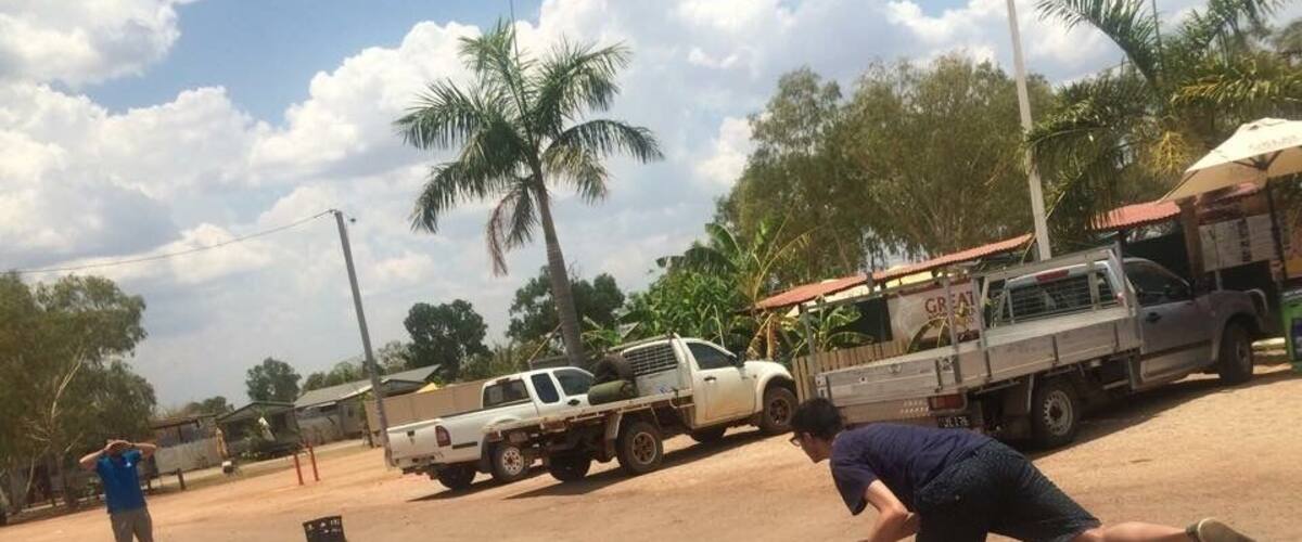 A dirt field with a white pickup truck, palm trees, and a building with a red roof in the background.