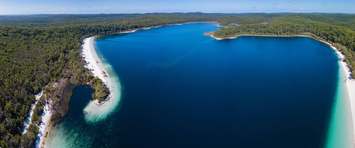 A large, clear blue lake surrounded by dense forest and white sandy beaches.