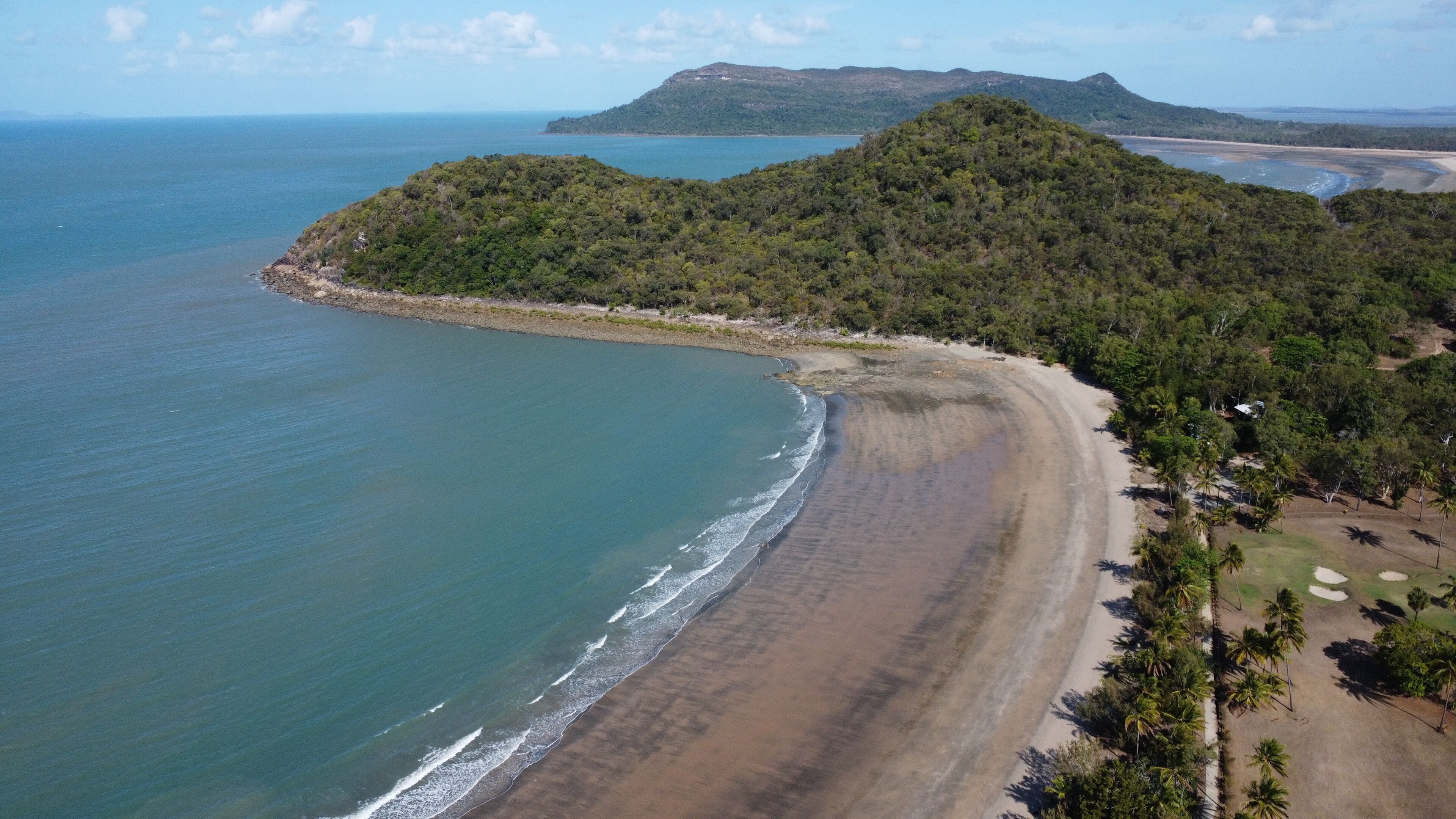 A coastal landscape with a sandy beach, a forested hill, and a distant mountain range.