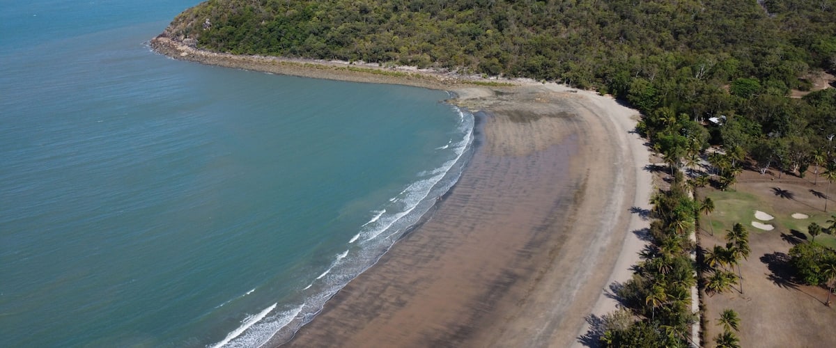 A coastal landscape with a sandy beach, a forested hill, and a distant mountain range.