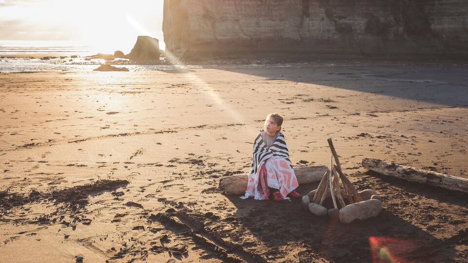 A person sitting on a beach with a blanket, surrounded by driftwood and rocks.