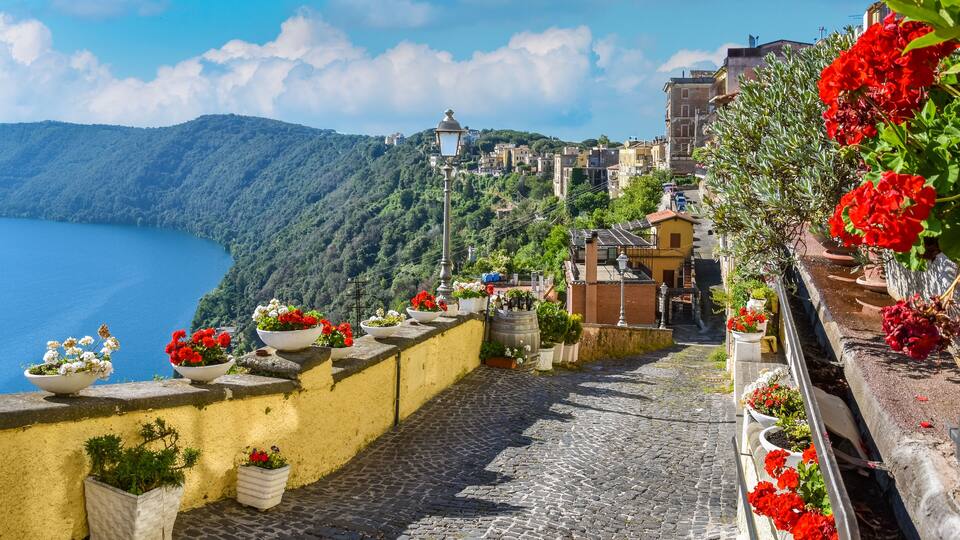 A cobblestone street with flower pots and a view of a lake and hills.